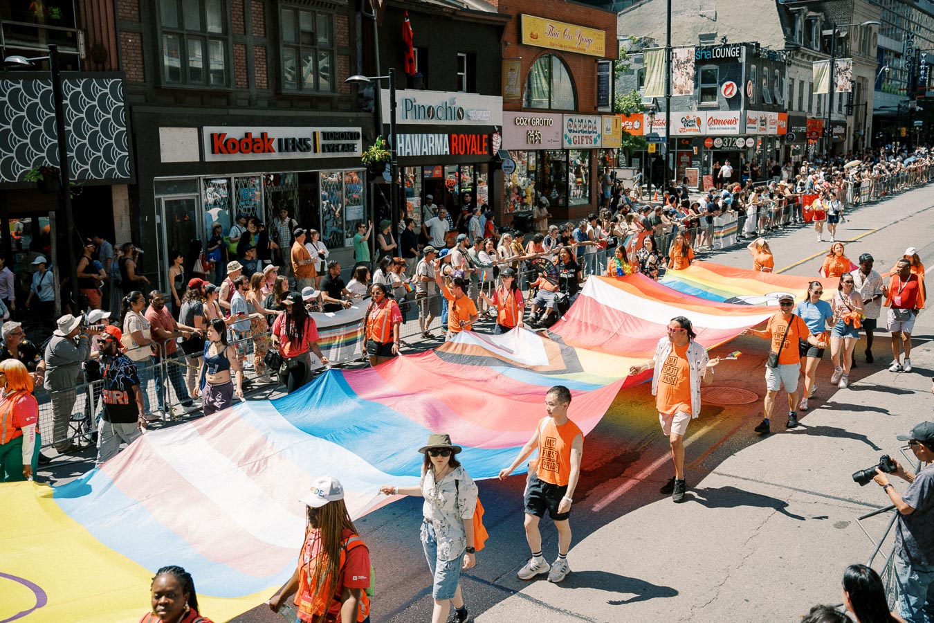 Participants in a pride parade carry a large rainbow flag down a bustling city street, with spectators lining the sidewalks.