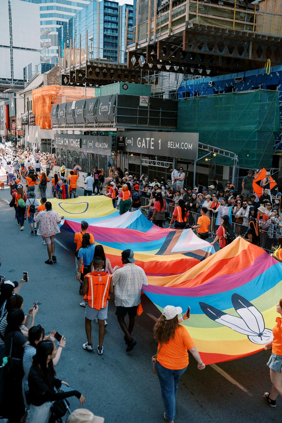 A colorful parade featuring a large rainbow flag being carried by participants down a city street, with spectators lining the sides.