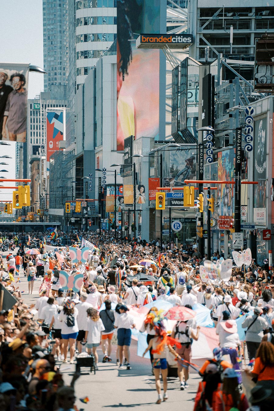 A lively parade takes place on a bustling city street, featuring participants with colorful flags and banners, surrounded by skyscrapers and digital billboards.