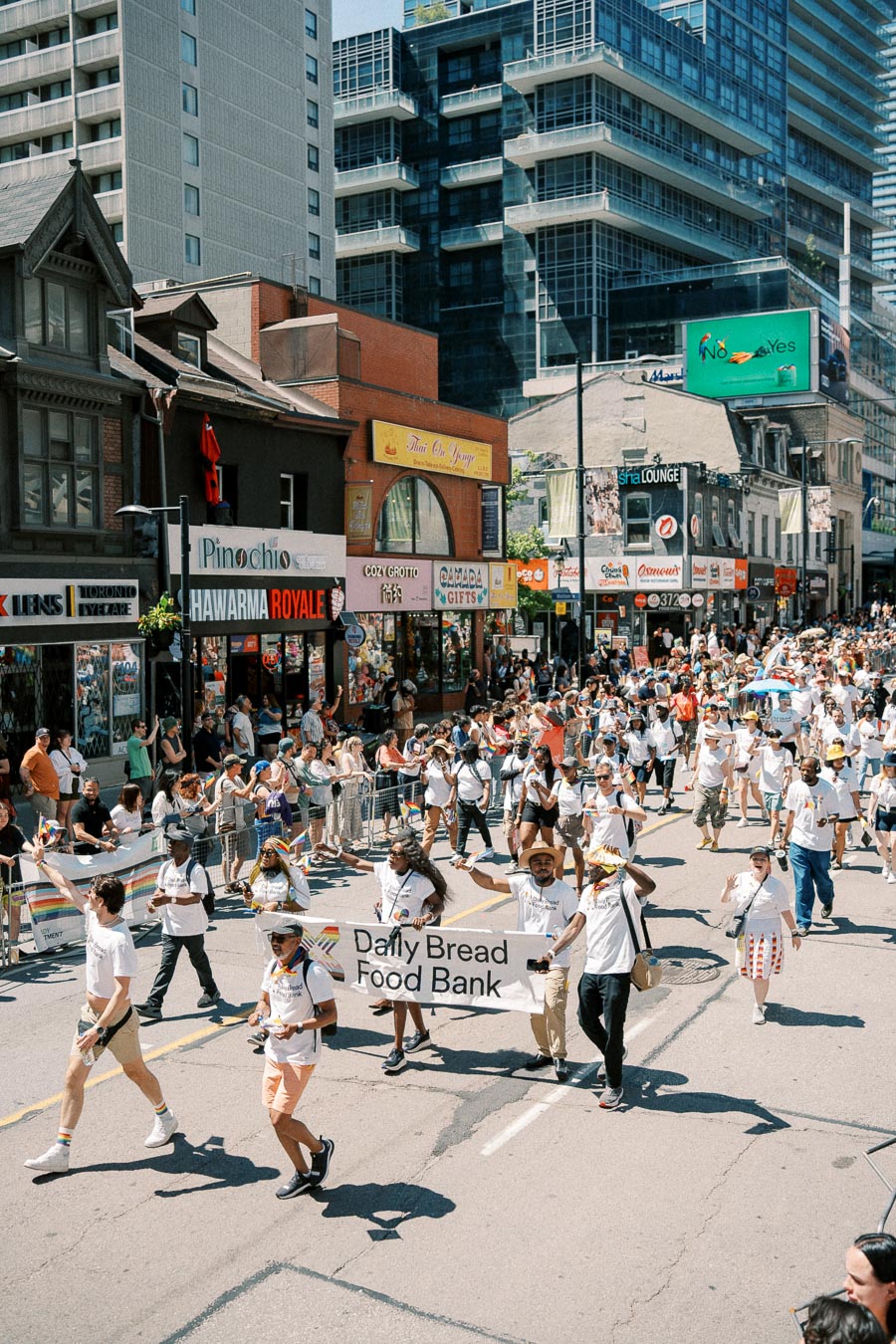 Participants in a parade walk down a city street, holding a banner for the Daily Bread Food Bank. The street is lined with shops and spectators.