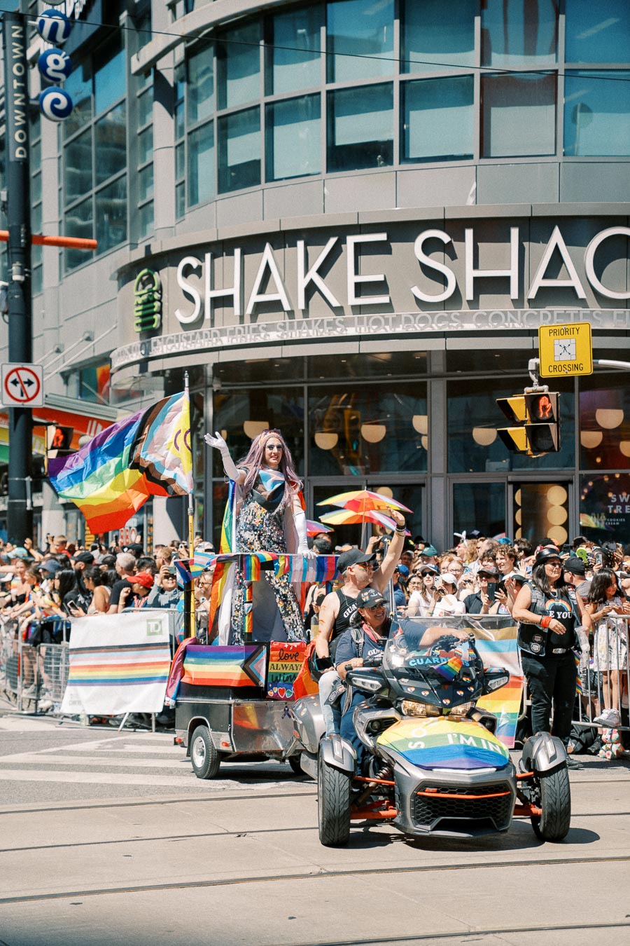A lively pride parade passes by a Shake Shack, featuring a colorful float and participants celebrating with rainbow flags.