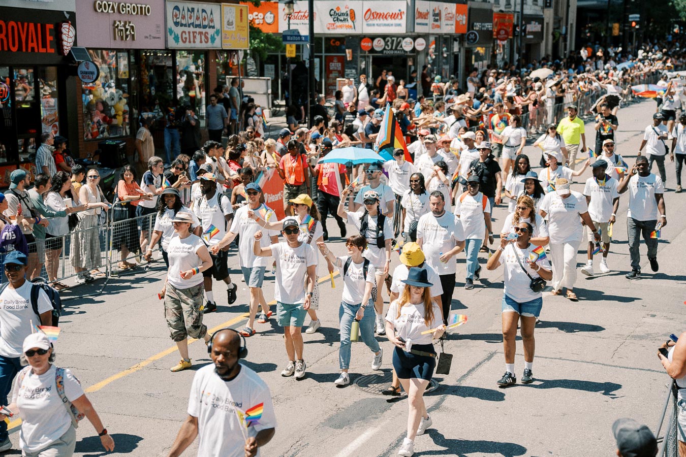 Participants in a parade march down a street, wearing "Daily Bread Food Bank" shirts and holding rainbow flags, while spectators watch from the sidelines.