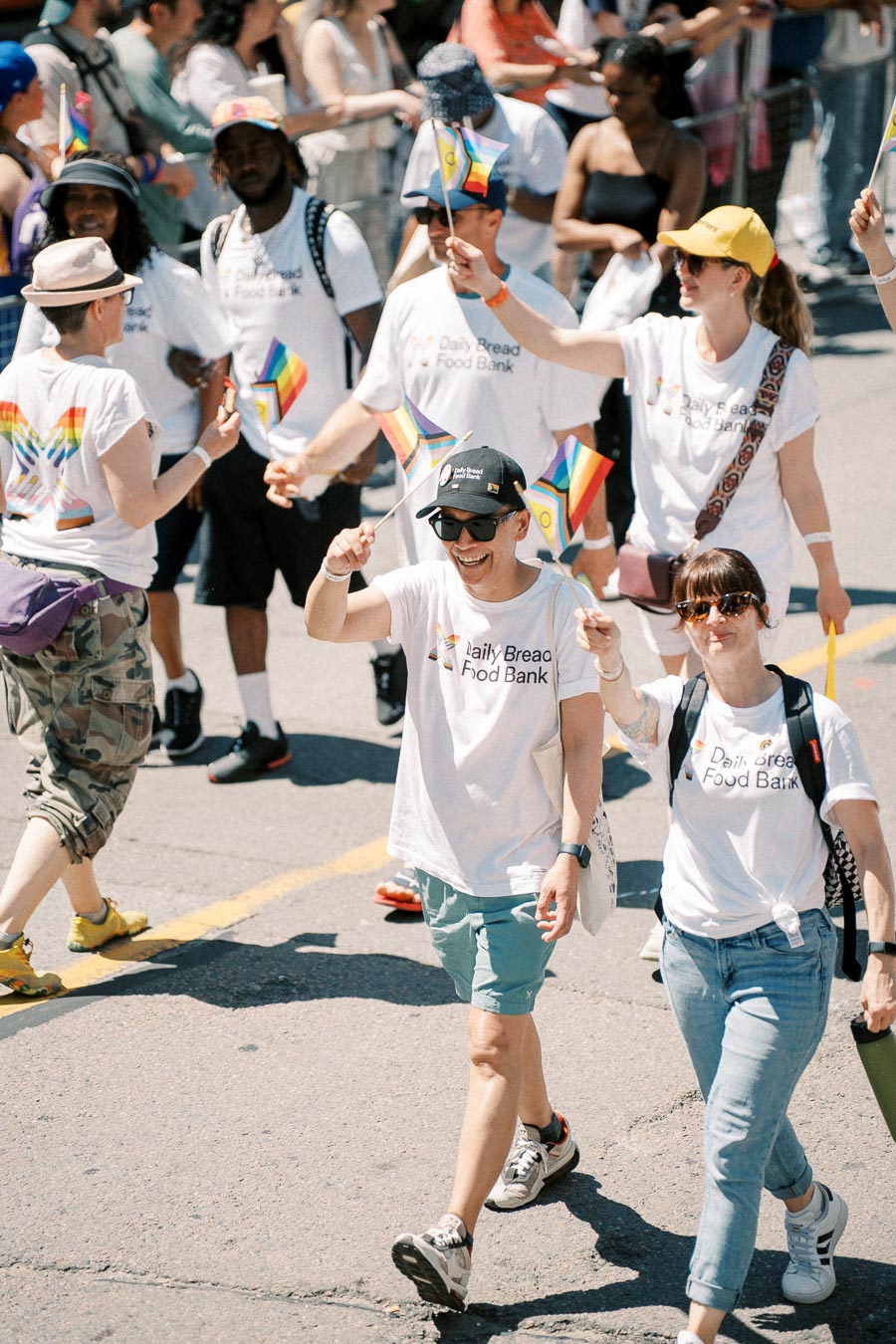Participants in a parade wearing "Daily Bread Food Bank" t-shirts and waving rainbow flags, celebrating diversity and community support.