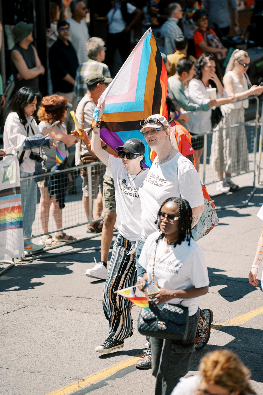 Participants in a pride parade hold a colorful pride flag, wearing 'Daily Bread Food Bank' shirts, surrounded by a crowd.