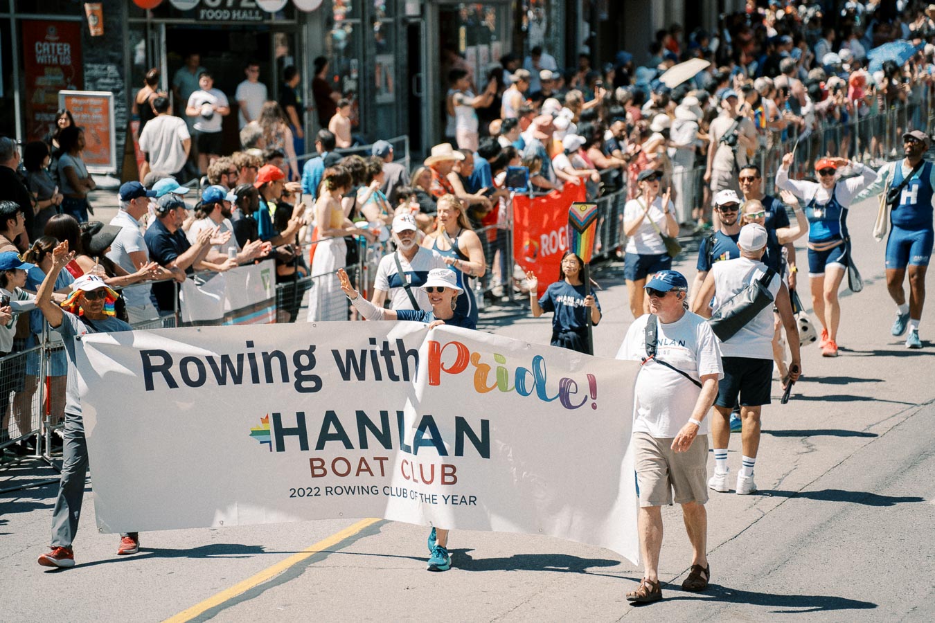 Participants from the Hanlan Boat Club march in a parade, holding a banner that reads 'Rowing with Pride! Hanlan Boat Club 2022 Rowing Club of the Year'. The event is lively, with a large crowd lining the street, celebrating diversity and inclusion.