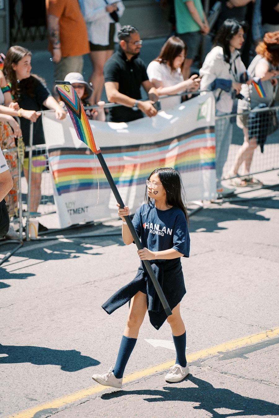 A participant in a parade carries a rainbow-themed paddle, symbolizing inclusivity and diversity, while spectators watch from the sidelines.