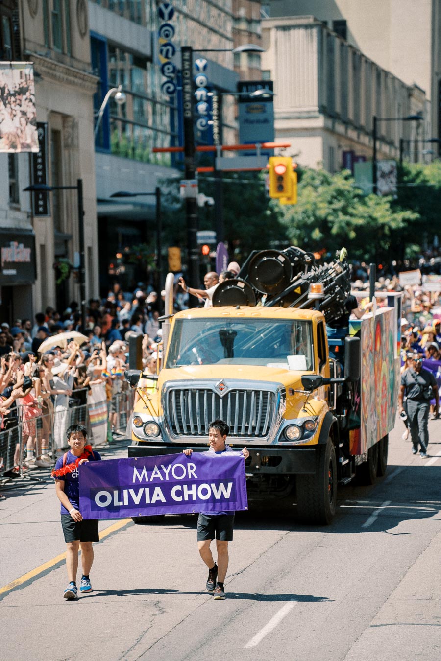 A parade featuring a yellow truck and participants holding a banner that reads 'Mayor Olivia Chow'. The event takes place on a city street lined with spectators.