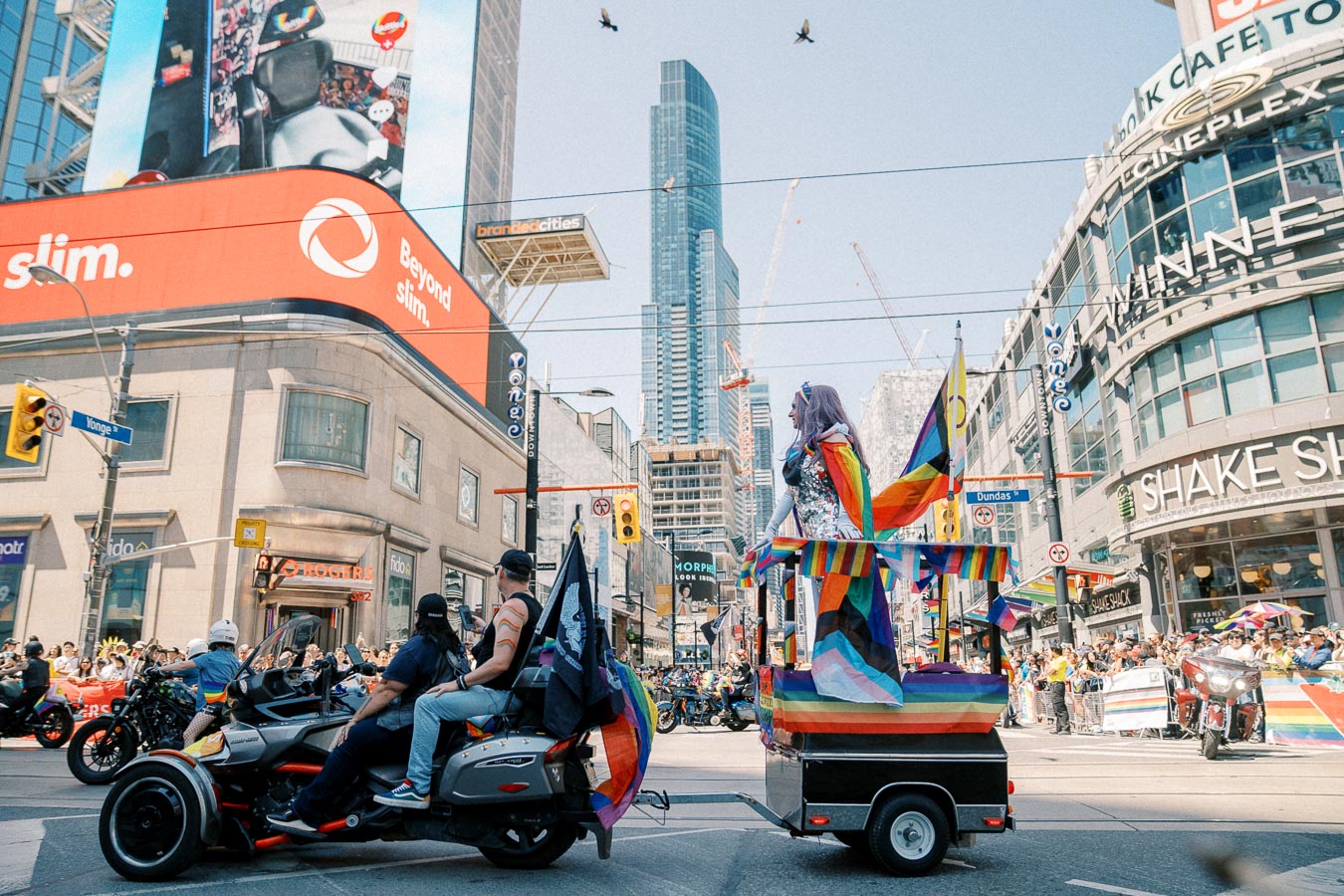 A lively pride parade in a bustling city, featuring a colorful float and participants on motorcycles, with spectators lining the streets.