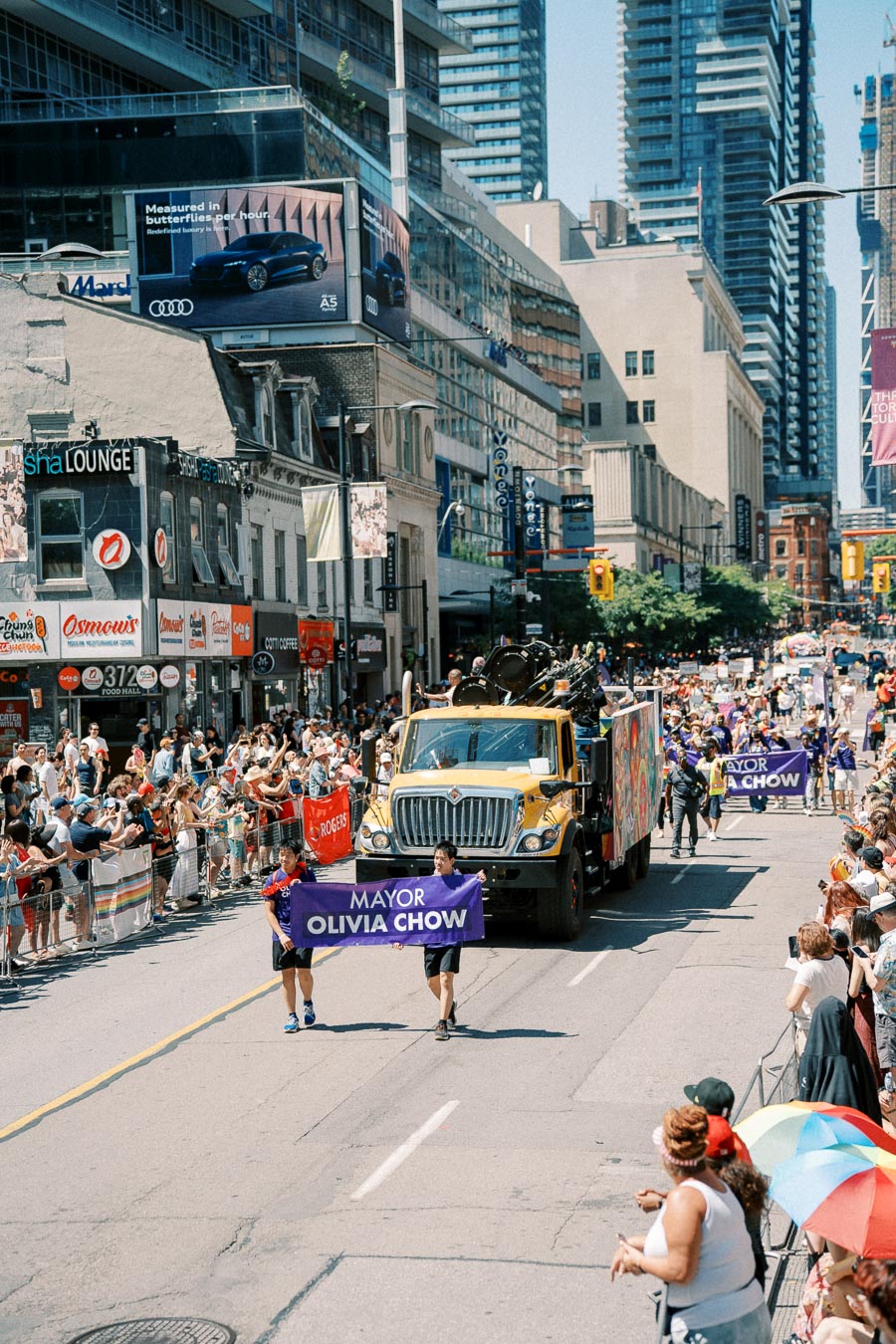 A vibrant city parade features a yellow truck and participants holding a banner for 'Mayor Olivia Chow'. Crowds line the street, enjoying the festive atmosphere.