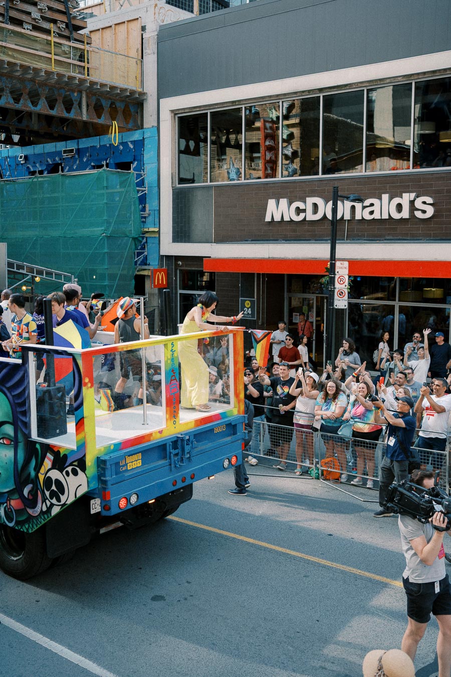 A vibrant parade float passes by a McDonald's during a street celebration, with people cheering and waving rainbow flags.