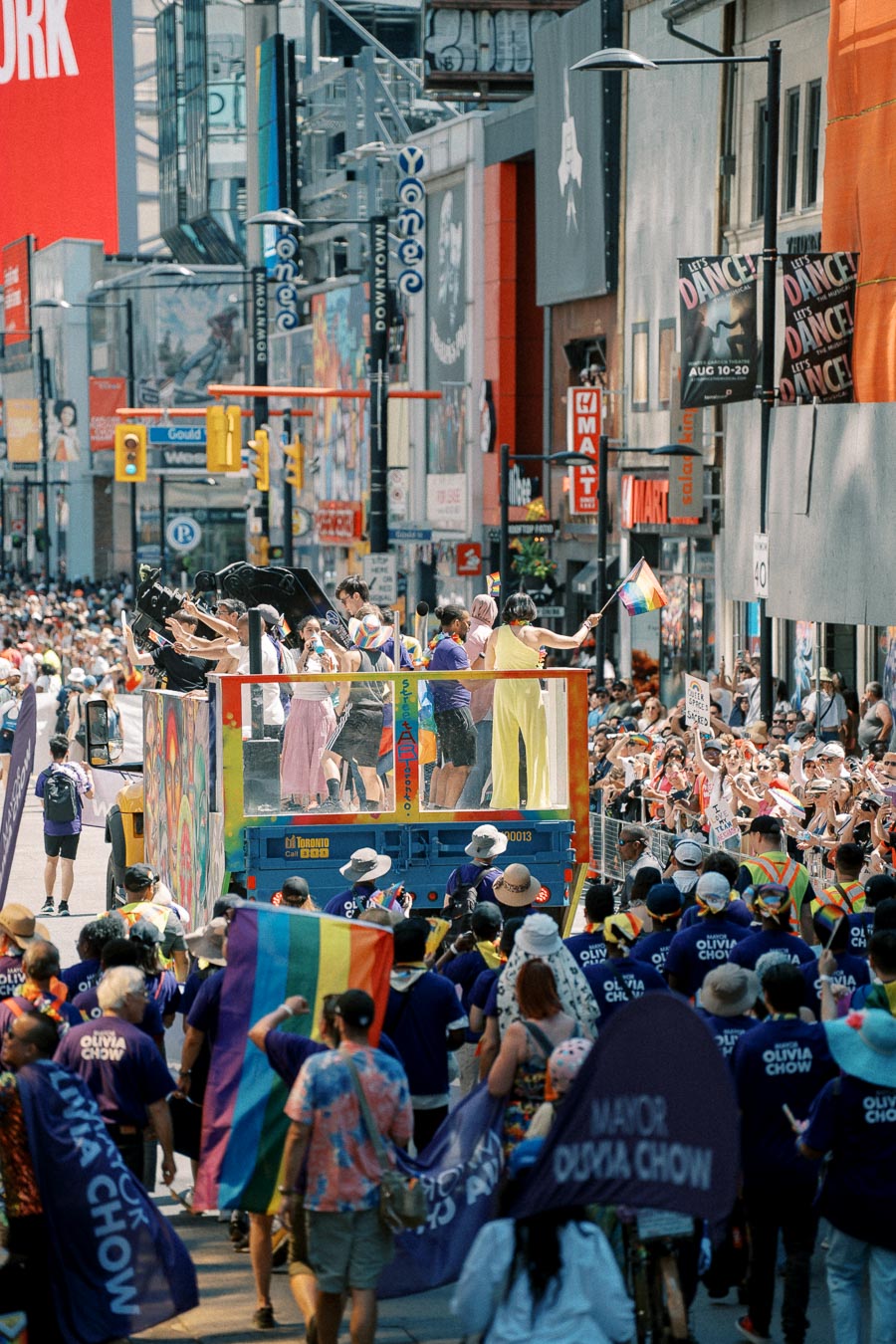 A lively parade scene with participants on a colorful float waving rainbow flags, surrounded by a cheering crowd on a bustling city street.