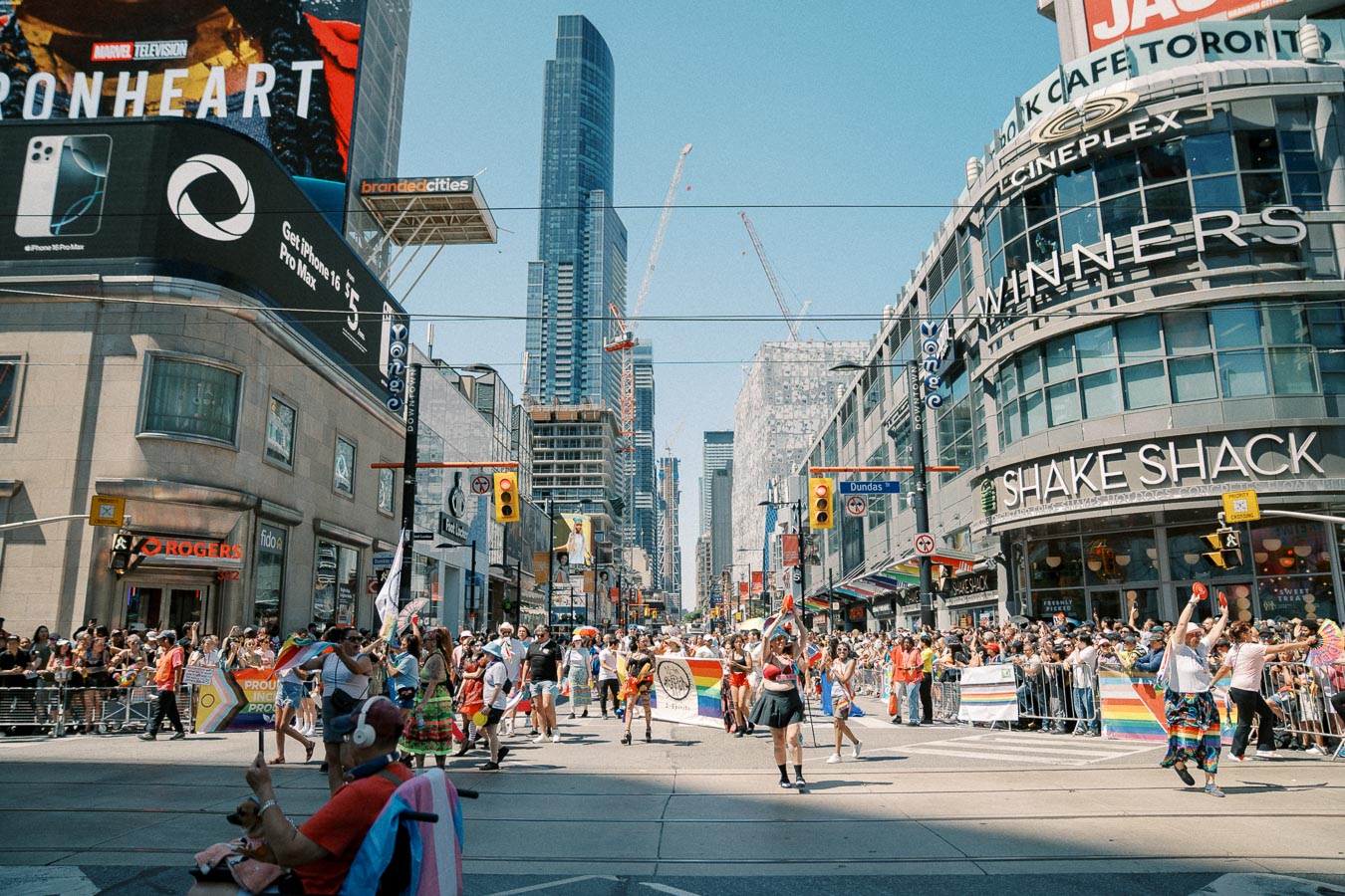 A lively parade in downtown Toronto, featuring participants with rainbow flags and banners, surrounded by cheering spectators and iconic city buildings.