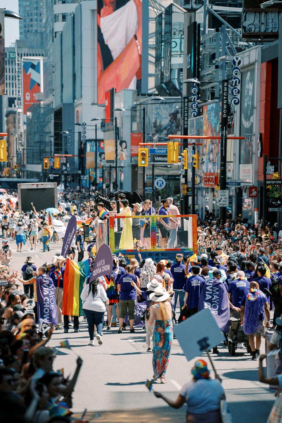 A lively parade featuring participants with rainbow flags and banners, drawing a large crowd in a bustling city street.