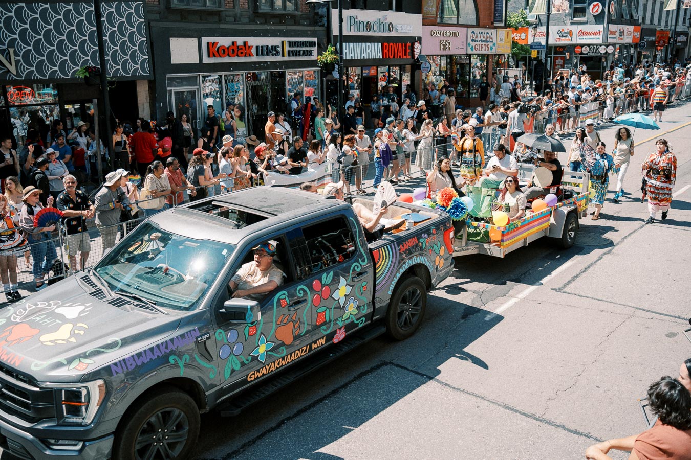 A vibrant parade features a decorated truck and trailer, with participants and spectators enjoying the event on a bustling city street.