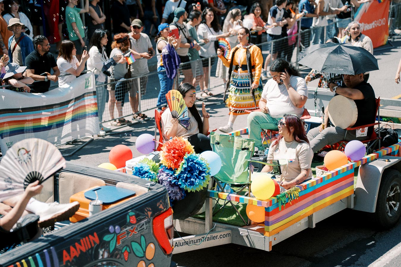 A colorful parade float adorned with rainbow decorations and balloons passes by a cheering crowd during a lively street parade.