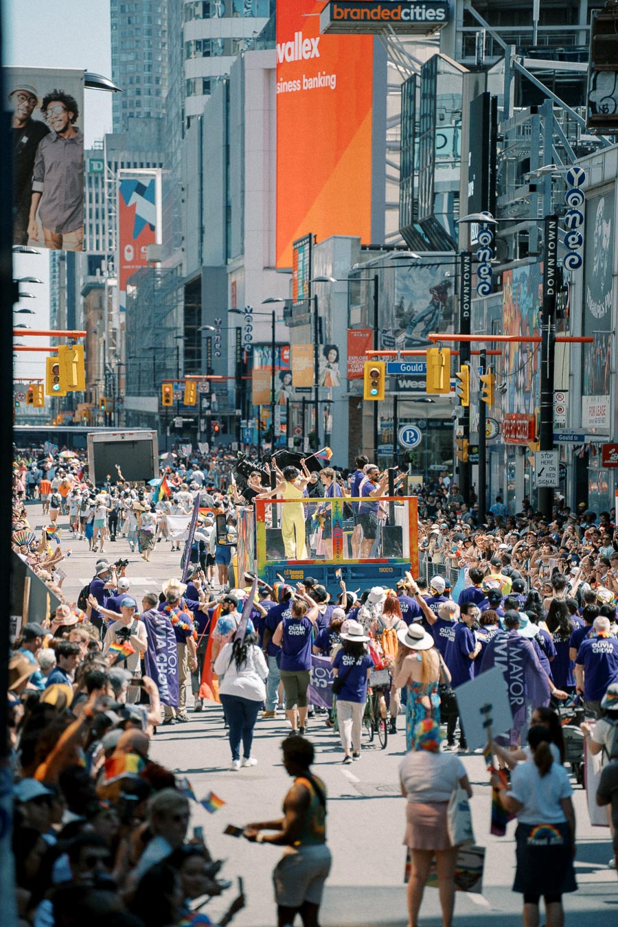 A lively parade takes place on a bustling city street, with participants in purple shirts and colorful flags, surrounded by skyscrapers and large billboards.