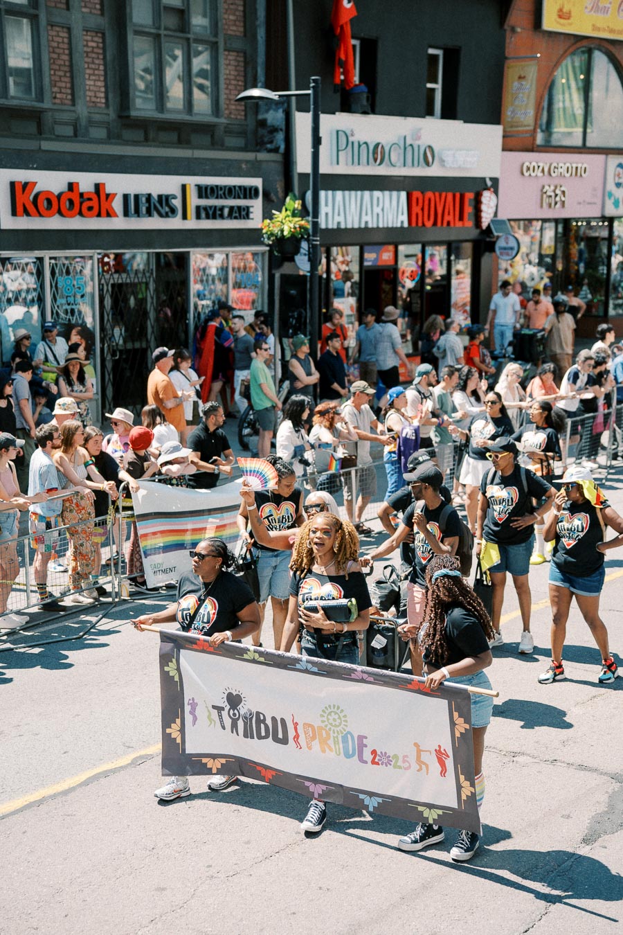 Participants march in a pride parade, holding a 'Pride 2025' banner, with spectators and shops in the background.