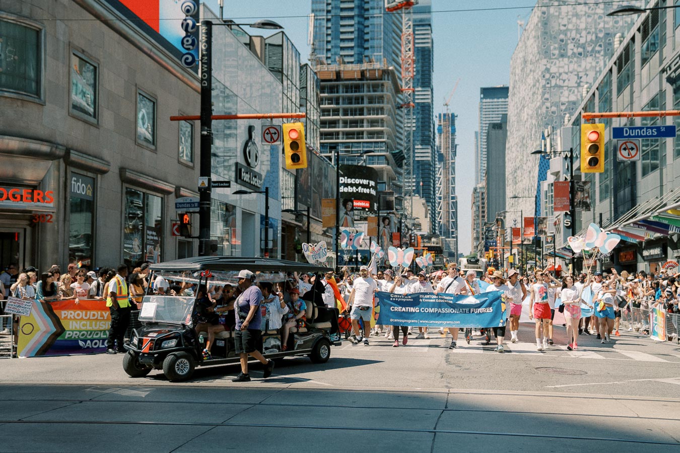 A lively parade takes place on Dundas Street, featuring participants with a banner and colorful balloons, surrounded by spectators and urban buildings.