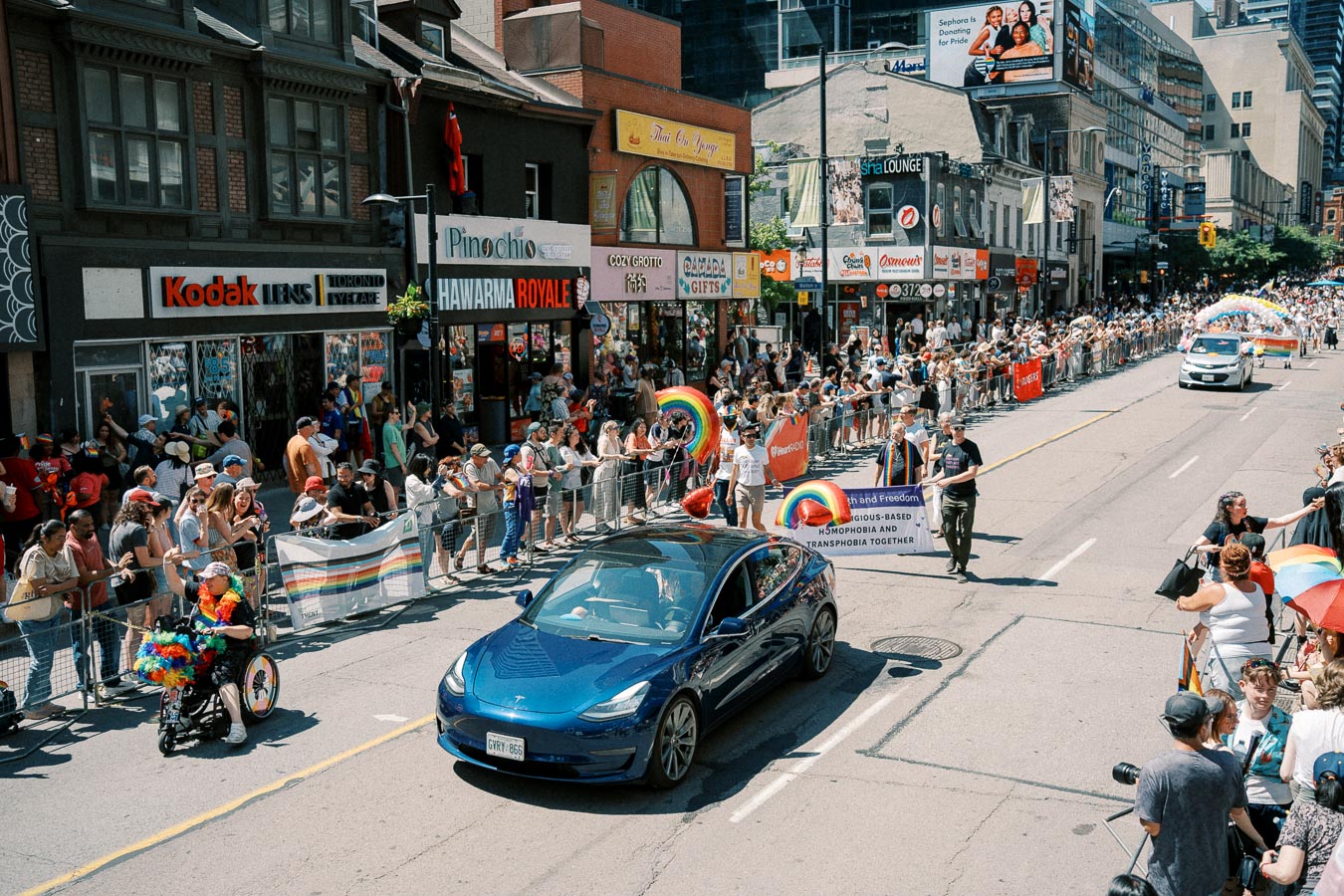 A lively street parade featuring a blue Tesla, rainbow flags, and a cheering crowd, celebrating diversity and inclusion.