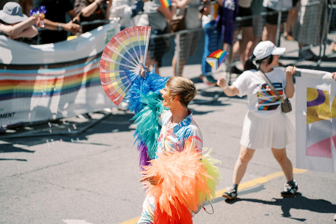 A person dressed in a vibrant, rainbow-colored outfit with feathered sleeves participates in a pride parade, holding a large rainbow fan. Spectators in the background wave rainbow flags, celebrating diversity and inclusion.