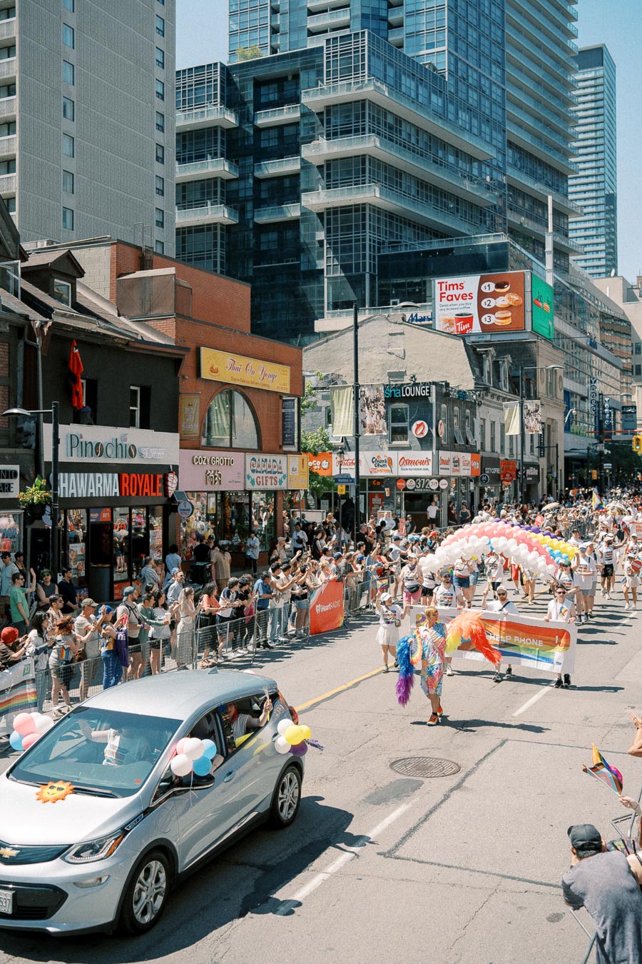 A lively parade takes place on a city street, featuring participants with colorful outfits and balloons, while spectators line the sidewalks. Tall buildings and various storefronts are visible in the background.