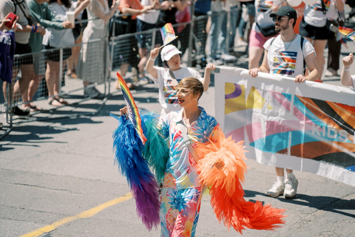 A vibrant participant at a pride parade, dressed in colorful attire and holding rainbow feather boas, engages with the crowd.