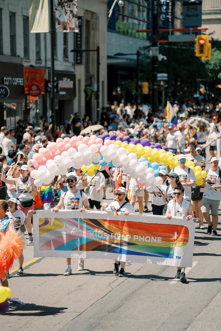 Participants in a pride parade march with a 'Kids Help Phone' banner, surrounded by colorful balloons and a festive crowd.