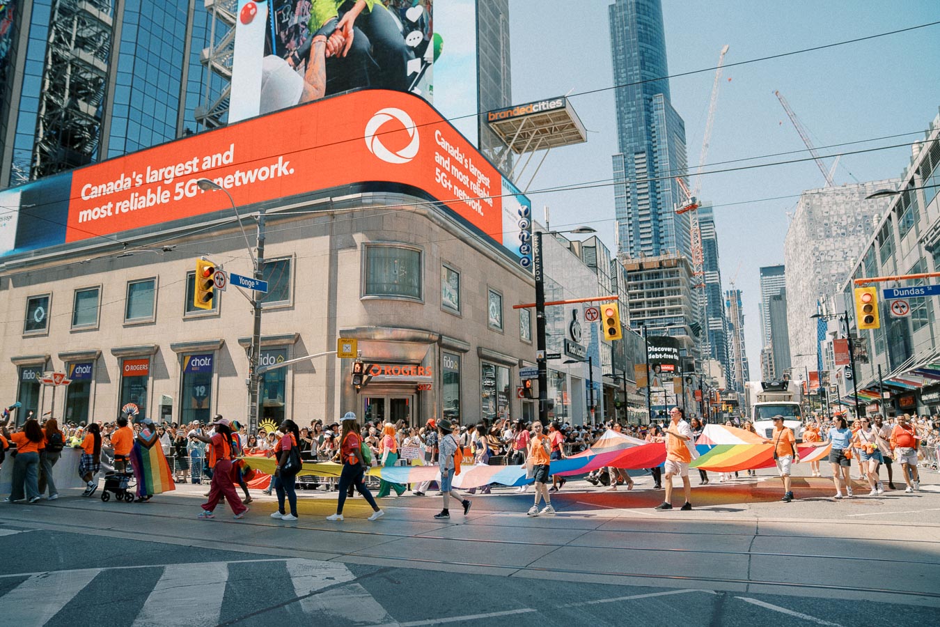 A colorful parade takes place at the intersection of Yonge and Dundas Streets in Toronto, featuring participants carrying a large rainbow flag. The scene is lively with a backdrop of skyscrapers and digital billboards.