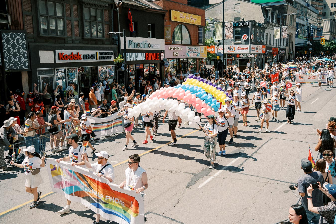 Participants march in a lively pride parade, carrying colorful banners and balloons, as spectators line the city street.