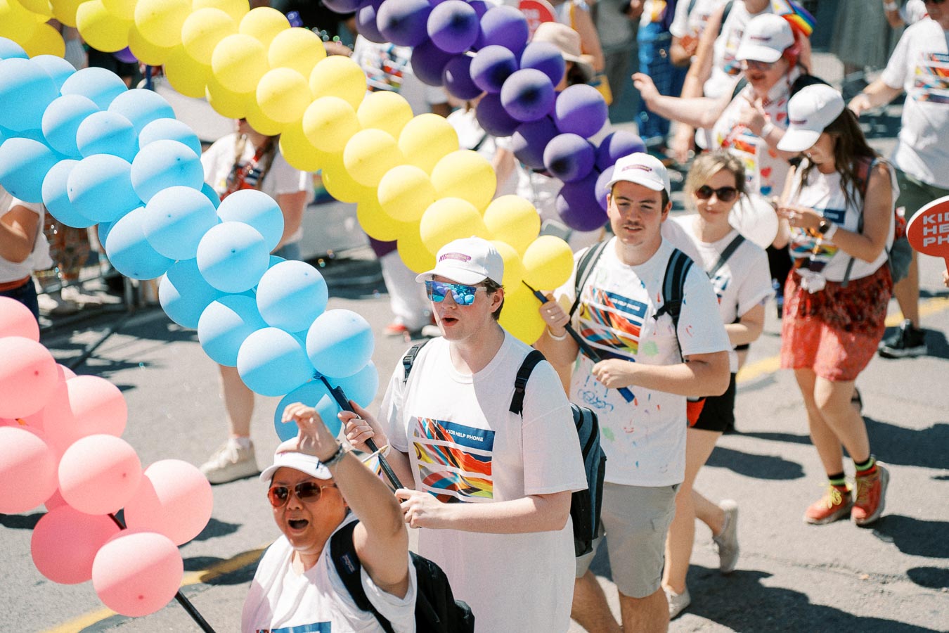 Participants in a parade hold colorful balloons, wearing white shirts and caps, celebrating diversity and inclusion.
