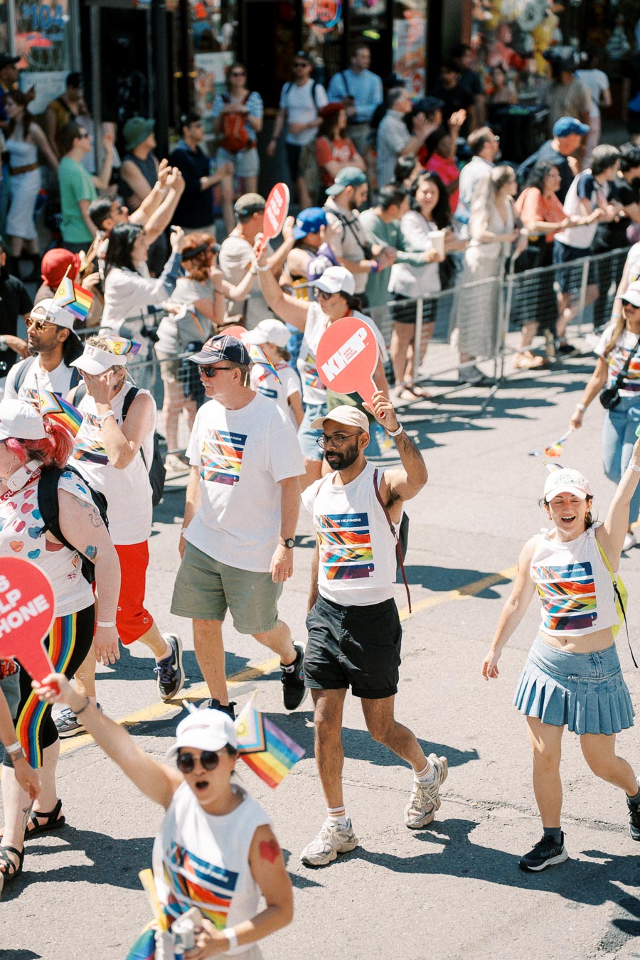 Participants in a pride parade walk down a street, wearing rainbow-themed attire and holding signs, while spectators watch from behind barriers.