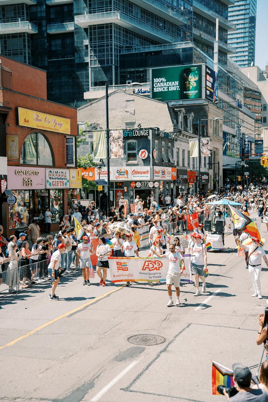 Participants in a pride parade march down a city street, holding a banner with the ADP logo. The street is lined with spectators and various storefronts, including Thai On Yonge and Canada Gifts. A Moosehead beer advertisement is visible on a nearby building.