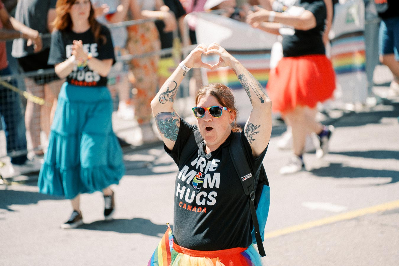 A participant at a pride parade enthusiastically makes a heart shape with their hands. They are wearing a "Free Mom Hugs Canada" shirt, rainbow sunglasses, and a colorful tutu, embodying the spirit of love and support.