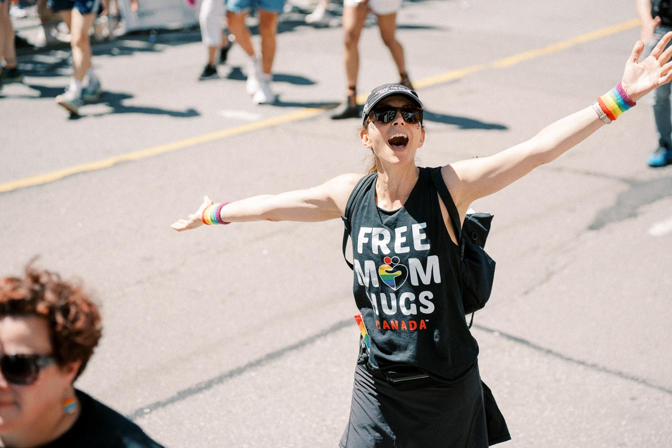 A joyful participant at a parade wearing a 'Free Mom Hugs Canada' shirt and rainbow accessories.