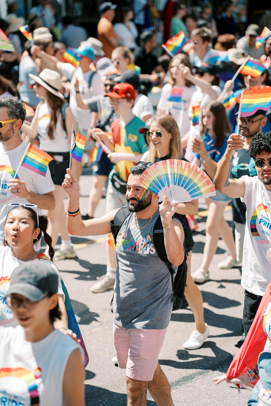 Participants celebrate at a pride parade, waving rainbow flags and fans, showcasing vibrant attire and joyful expressions.