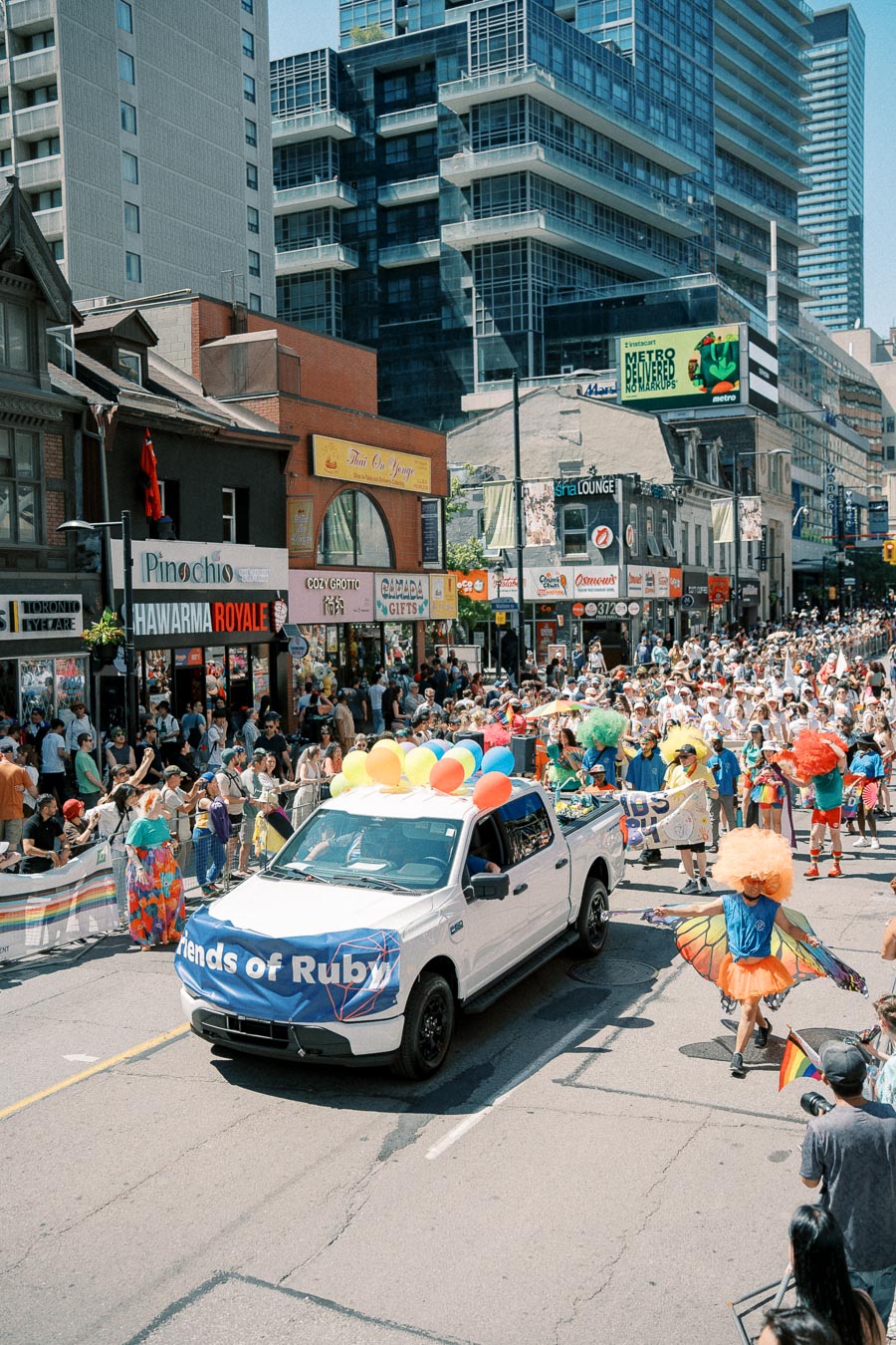 A vibrant parade featuring a white truck with a 'Friends of Ruby' banner, surrounded by people in colorful costumes and a cheering crowd.