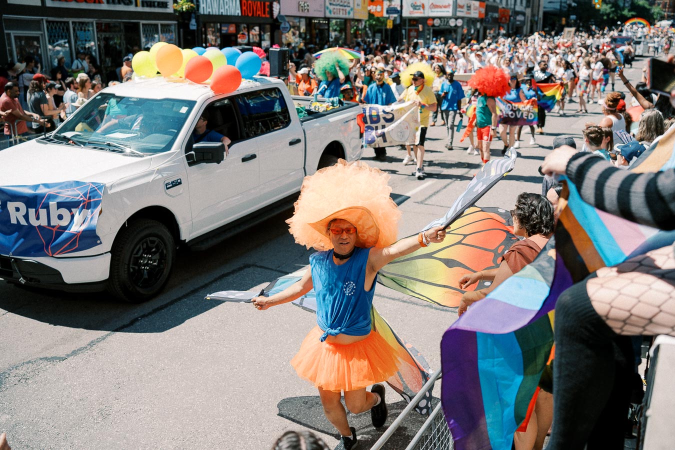 A lively scene from a pride parade featuring a person in an orange tutu and hat, a white truck adorned with colorful balloons, and a crowd waving rainbow flags.