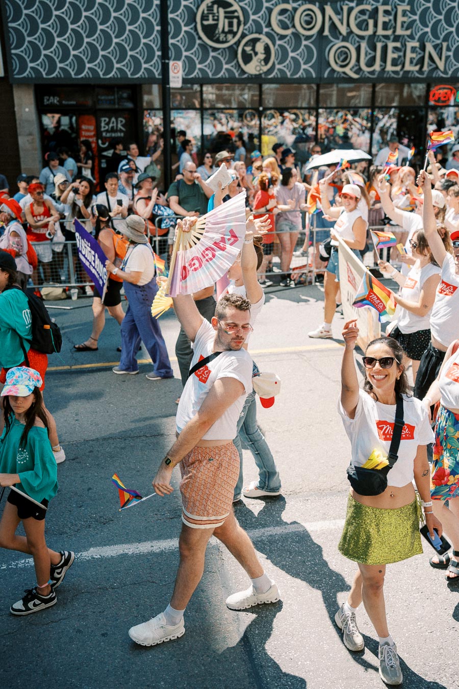 Participants in a vibrant pride parade wave rainbow flags and fans, celebrating diversity and inclusion. Spectators line the sidewalk, enjoying the festive atmosphere.