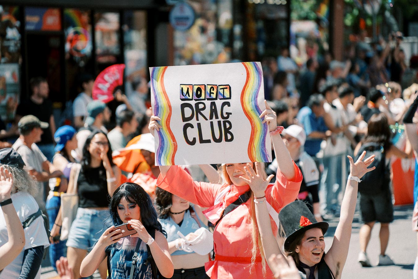 A participant holds a colorful 'UofT Drag Club' sign at a lively outdoor event, surrounded by a diverse crowd.