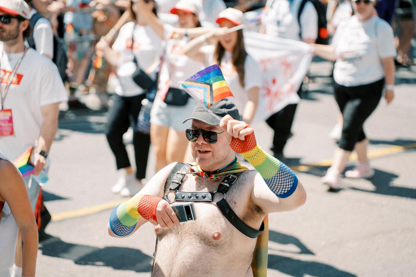 A participant in a pride parade wearing colorful rainbow accessories and holding a pride flag, surrounded by other marchers.