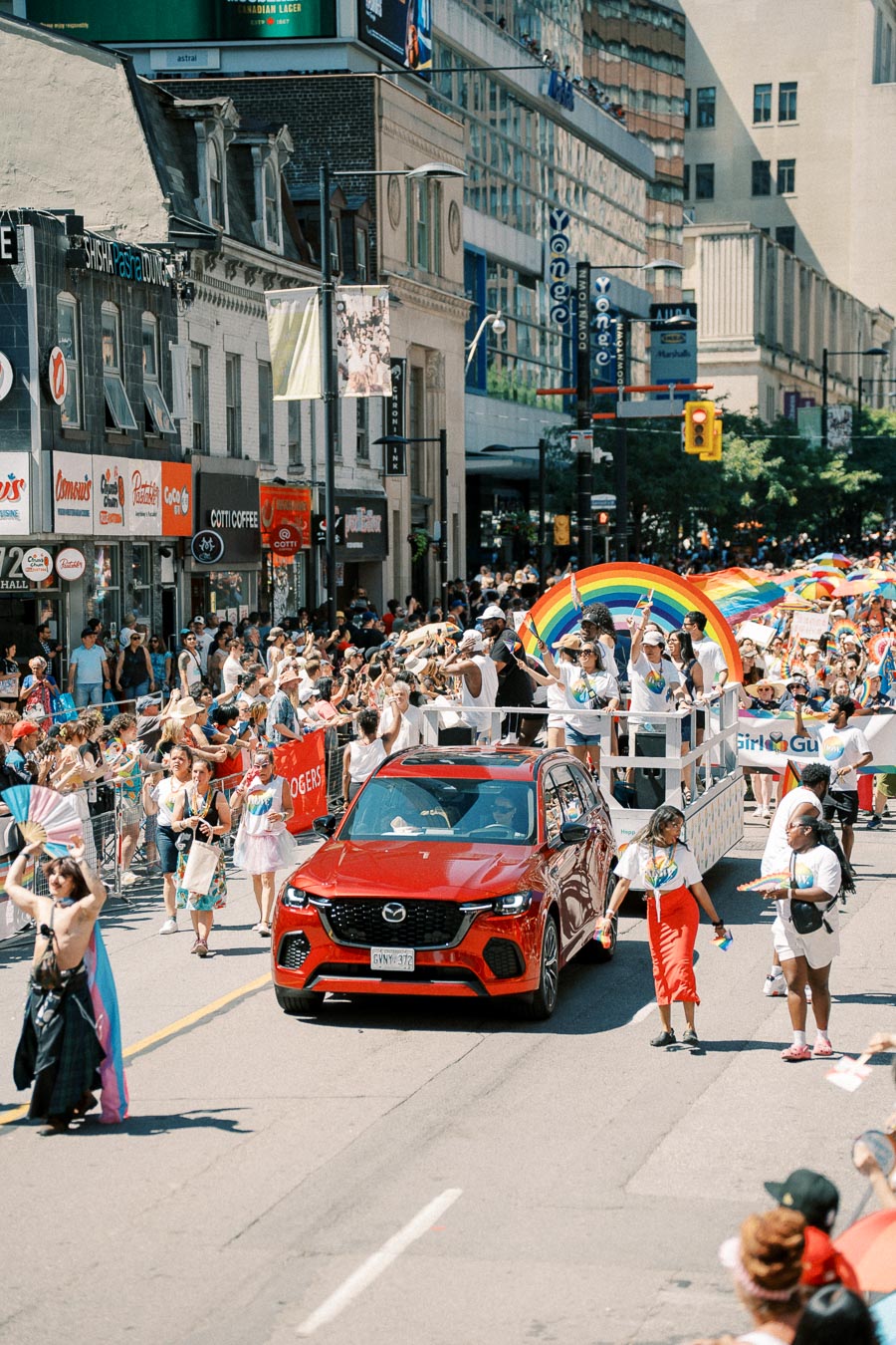 A lively parade scene featuring a red Mazda car and a float with a rainbow arch, surrounded by participants and spectators celebrating with rainbow flags.