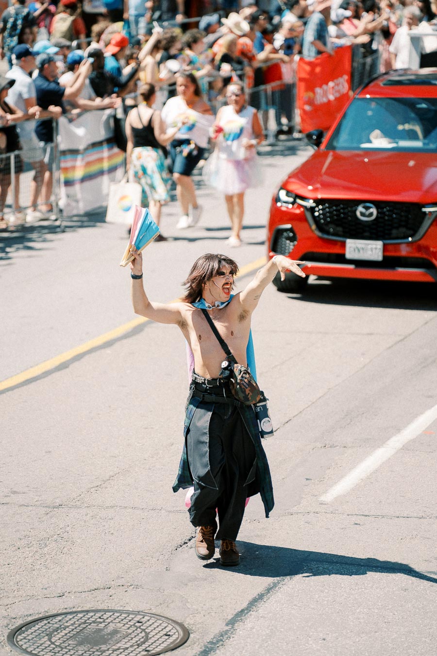 A lively scene from a pride parade featuring a person joyfully waving colorful fans, surrounded by a cheering crowd and a red Mazda car.