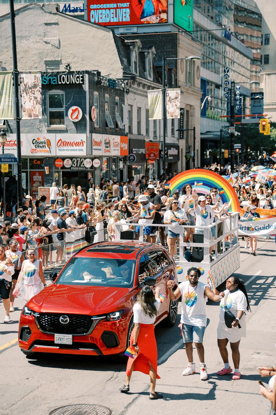 A lively parade scene featuring a red Mazda SUV and a float with a rainbow arch. Participants are celebrating with colorful attire and flags, creating a festive atmosphere.