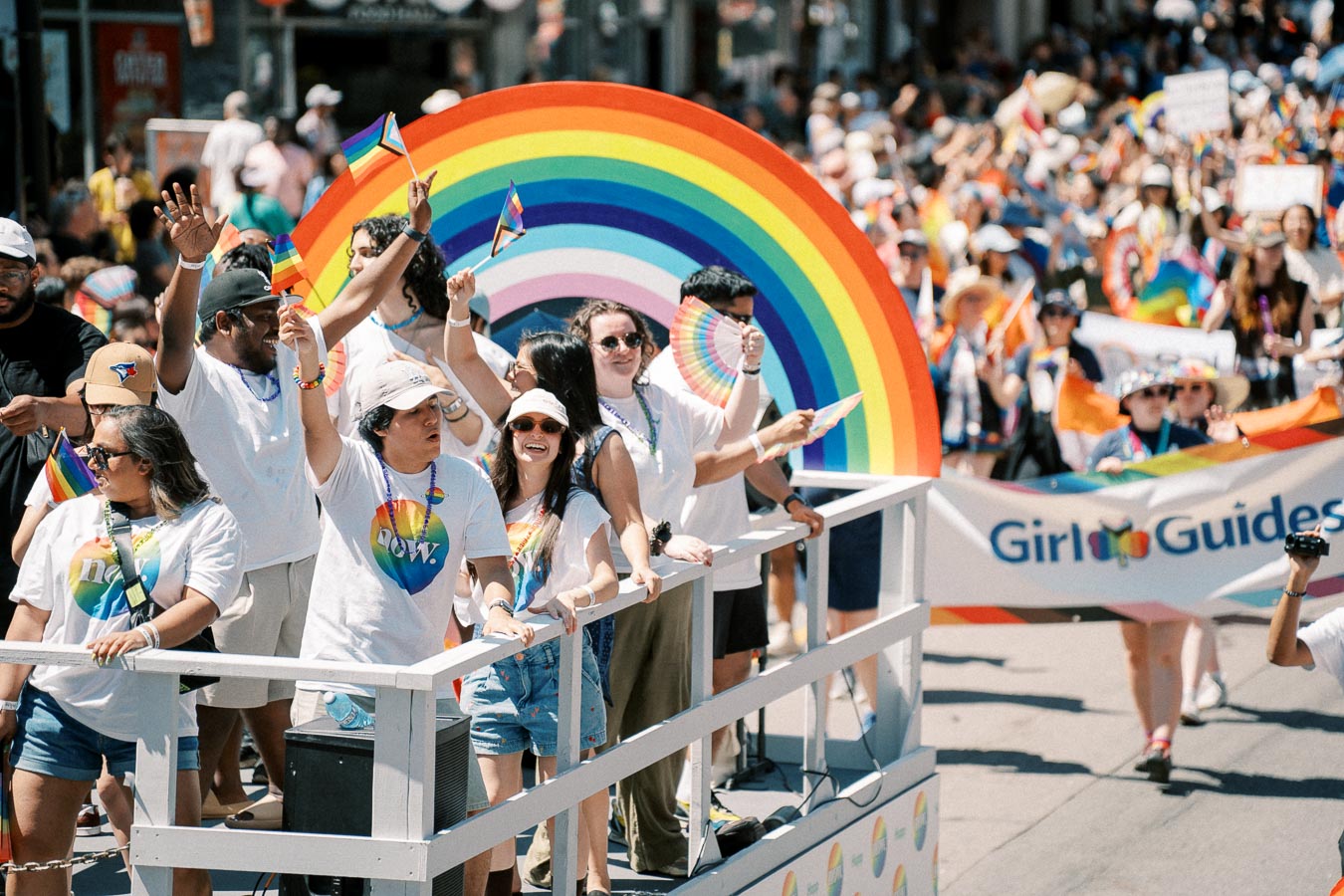 Participants celebrate at a Pride parade, waving rainbow flags and wearing themed attire, with a large rainbow arch and a 'Girl Guides' banner in the background.