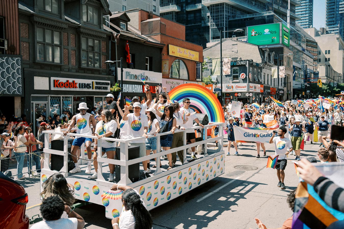 A lively scene from a pride parade featuring a colorful float with a rainbow and participants wearing matching shirts, surrounded by a cheering crowd.