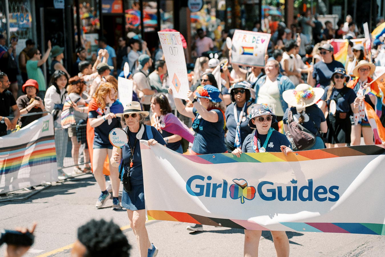 Participants in a parade hold a 'Girl Guides' banner, celebrating LGBTQ+ pride with colorful signs and attire.