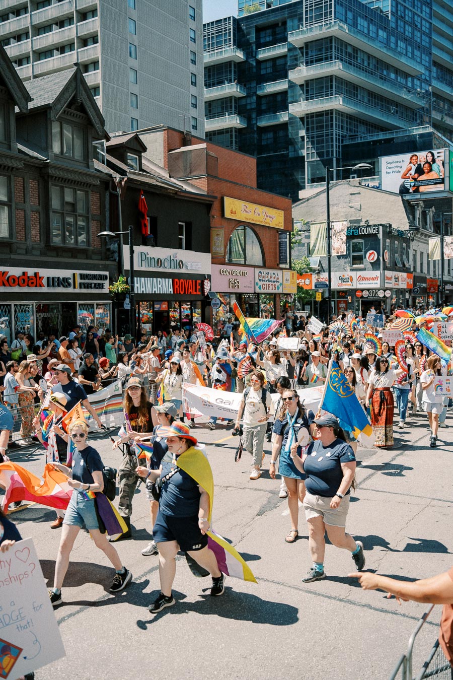 Participants march in a colorful pride parade, carrying rainbow flags and banners, along a bustling city street lined with shops and modern buildings.