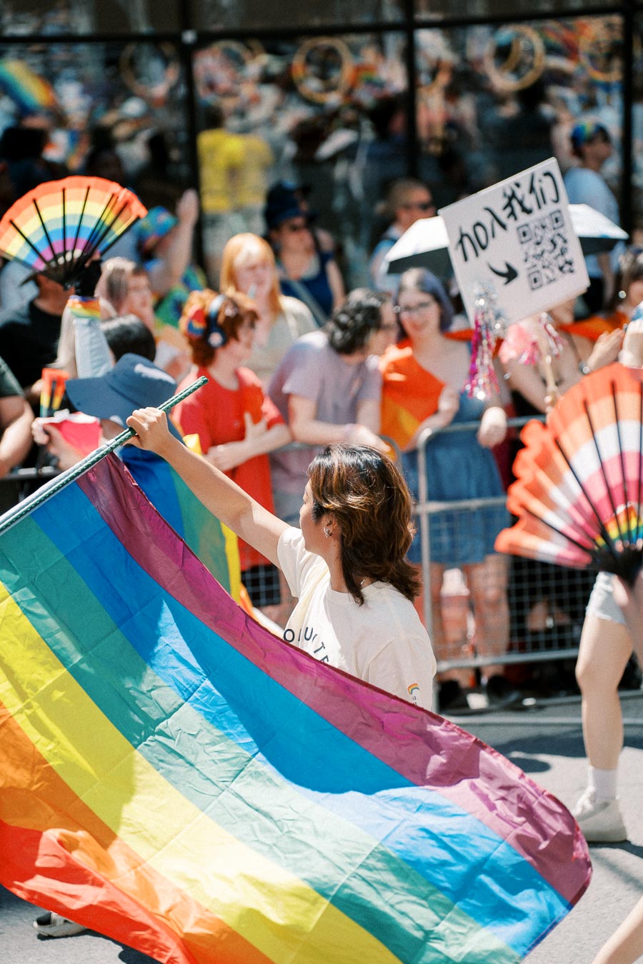 A vibrant scene from a pride parade featuring a person holding a rainbow flag, surrounded by a lively crowd with colorful fans and signs.