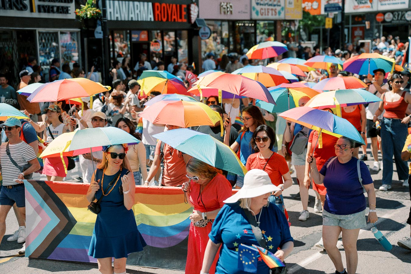 Participants in a pride parade hold colorful rainbow umbrellas and a pride flag as they walk down a bustling street.