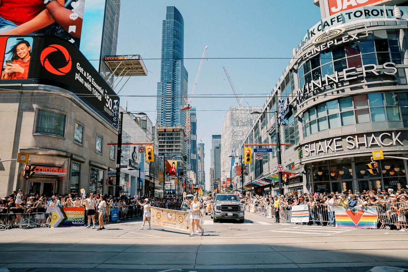 A vibrant parade in a bustling city center, featuring participants marching with banners and rainbow flags, suggesting a pride celebration. The scene is set against a backdrop of tall buildings and popular establishments like Shake Shack and Cineplex.